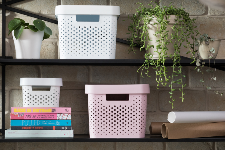 Three storage baskets in various sizes are sat on two shelves next to plants and books in a home office.