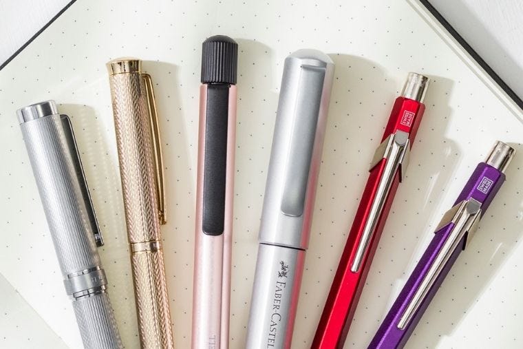 A selection of premium pens are lined up on an open page of a dotted notebook.