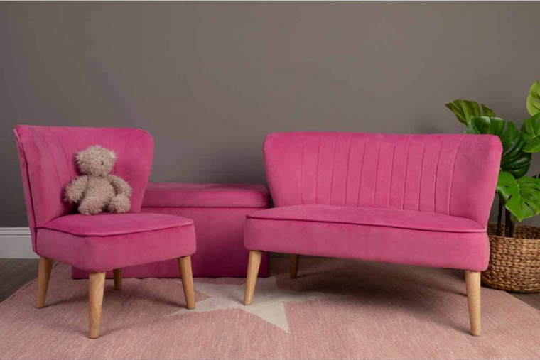 A pink velvet kids ottomon sits between two pieces of pink velvet kids furniture in a bedroom.